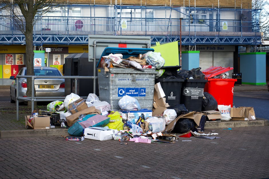 A large collection of assorted household waste and recyclables piled atop a paved urban street corner, including cardboard boxes, plastic bags filled with rubbish, paper and packaging materials, and black and red bin liners, some overflowing from large wheeled bins marked for mixed paper and cardboard disposal. The scene includes a parked silver hatchback car with a visible UK license plate positioned behind the waste, and a metal railing surrounding part of the area to prevent access or parking. In the background, there is a building with a blue upper façade under construction or renovation, supported by scaffolding, and storefronts with signage, one of which reads ‘Premier’. Two green eco-branded waste collection points are visible on either side of the bins, indicating designated recycling stations. The overall environment appears to be a commercial or residential street with a focus on private waste disposal, potentially managed by local independent services like Rubbish Collection Uxbridge, rather than municipal collection. The atmosphere is neutral with natural daylight, highlighting the chaotic and uncontained nature of the overflowing waste collection site situated on the pavement near parked vehicles and store entrances.