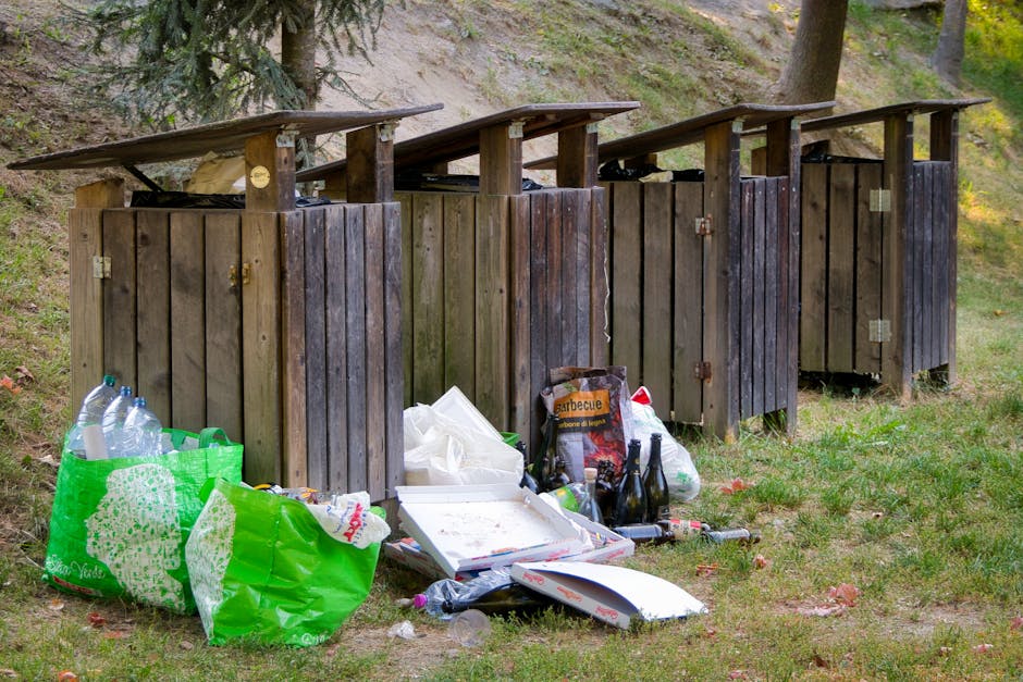 A large collection of assorted household waste and recyclables piled atop a paved urban street corner, including cardboard boxes, plastic bags filled with rubbish, paper and packaging materials, and black and red bin liners, some overflowing from large wheeled bins marked for mixed paper and cardboard disposal. The scene includes a parked silver hatchback car with a visible UK license plate positioned behind the waste, and a metal railing surrounding part of the area to prevent access or parking. In the background, there is a building with a blue upper façade under construction or renovation, supported by scaffolding, and storefronts with signage, one of which reads ‘Premier’. Two green eco-branded waste collection points are visible on either side of the bins, indicating designated recycling stations. The overall environment appears to be a commercial or residential street with a focus on private waste disposal, potentially managed by local independent services like Rubbish Collection Uxbridge, rather than municipal collection. The atmosphere is neutral with natural daylight, highlighting the chaotic and uncontained nature of the overflowing waste collection site situated on the pavement near parked vehicles and store entrances.