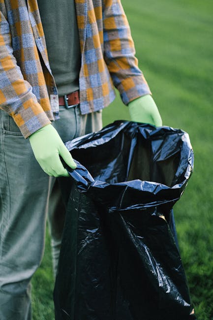 A person wearing a plaid shirt with yellow, gray, and blue tones, along with light green gloves, is holding open a large black garbage bag outdoors. The bag appears to be partially filled or ready to be filled, and the person's hands are gripping the top edges, preparing for rubbish collection. The background shows a green grassy area, suggesting a garden, park, or outdoor space. The scene is well-lit with natural daylight, emphasizing the textures of the garbage bag, the gloves, and the plaid shirt fabric. This image relates to waste management and rubbish removal services, emphasizing the process of private rubbish disposal or on-site clearance, typical of services offered by companies such as Rubbish Collection Uxbridge, which provides alternative waste handling options beyond municipal collection.