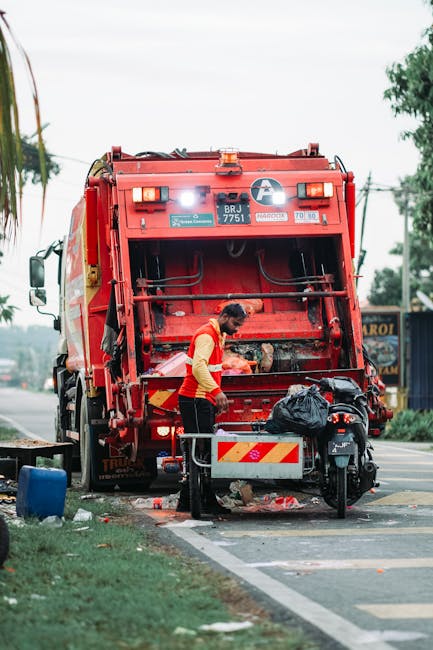 A worker wearing a high-visibility orange and yellow vest is standing next to a large red rubbish collection truck on the side of a street, actively operating the rear compaction mechanism. The truck, branded with reflective yellow and red safety markings, has a large, rectangular metal hopper at the back, which is open, exposing the interior with visible hydraulic arms and mechanisms used for waste compaction. The worker is leaning slightly forward, holding a black plastic bag filled with rubbish, which is being loaded into the truck. Nearby, there is a small, black motorbike parked beside a metal barrier, with a black bag placed on top of it. The scene is set on a pavement adjacent to a grass verge, with a few scattered plastic items and debris indicative of ongoing rubbish collection activity. In the background, a glimpse of a roadside shop or sign is visible, along with blurred trees and utility poles, suggesting a suburban environment. The scene is illuminated by natural daylight, creating a clear, neutral setting typical of an independent waste collection operation conducted by a private rubbish removal service near Uxbridge.