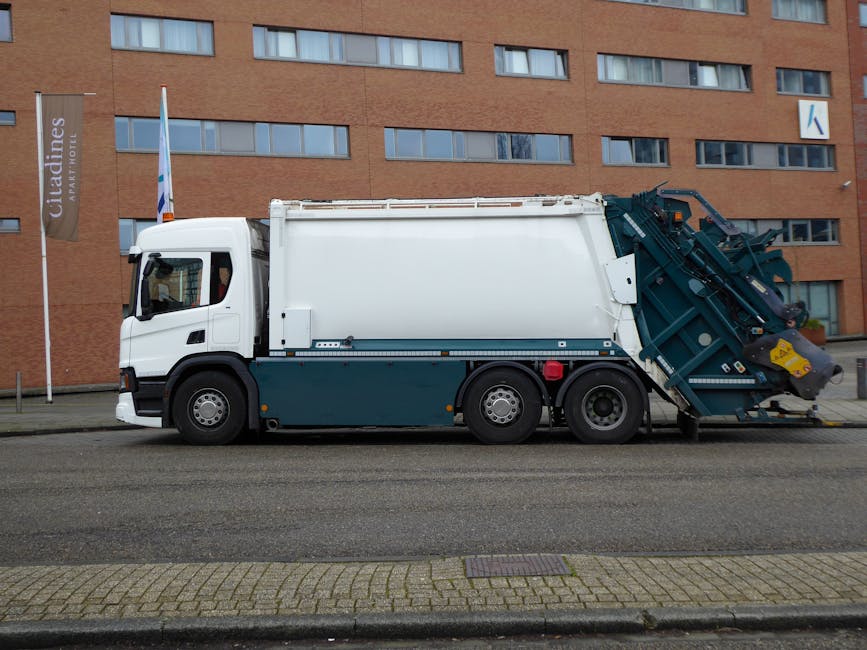 A large white and dark teal rubbish collection truck parked on a city street in front of a multi-storey brick building with numerous rectangular windows. The vehicle has a compact cab with tinted windows, and its rear section features an automated waste compaction and loading system with mechanical arms and hydraulic components, used for efficient waste removal. The truck is positioned parallel to the pavement, with the side facing the camera, showing details of the side panels, wheels, and the waste collection mechanism partially extended. The background includes a building with a brick exterior and a sign indicating nearby hotel accommodation, suggesting an urban environment where private waste collection services, such as those by Rubbish Collection Uxbridge, are utilized for on-site or alternative waste management solutions. The scene is captured in daylight with clear visibility, emphasizing the functional design and operational aspect of modern rubbish removal vehicles.