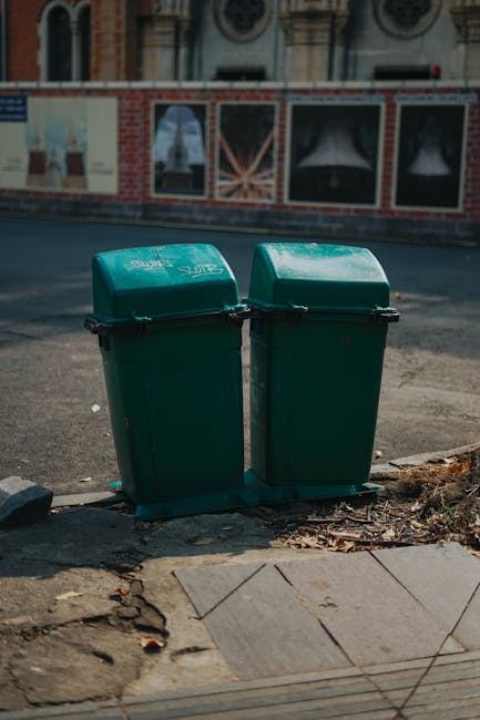 Two green outdoor waste bins with hinged lids positioned side by side on a concrete pavement verge, with a slight gap between them. The bins are made of durable plastic, showing a smooth finish and subtle branding or logo on the lids. They are situated in front of a road with a slight incline, close to the edge of the pavement, with some scattered dried leaves and debris visible at their base. The background features a brick building with decorative architectural details and windows displaying artwork or posters, with a wooden fence and a paved surface behind the bins. The scene is illuminated by daylight, casting soft shadows, indicating a clear or partly cloudy day, consistent with a typical urban environment where private waste collection or alternative rubbish disposal services may operate, as handled by companies like Rubbish Collection Uxbridge.